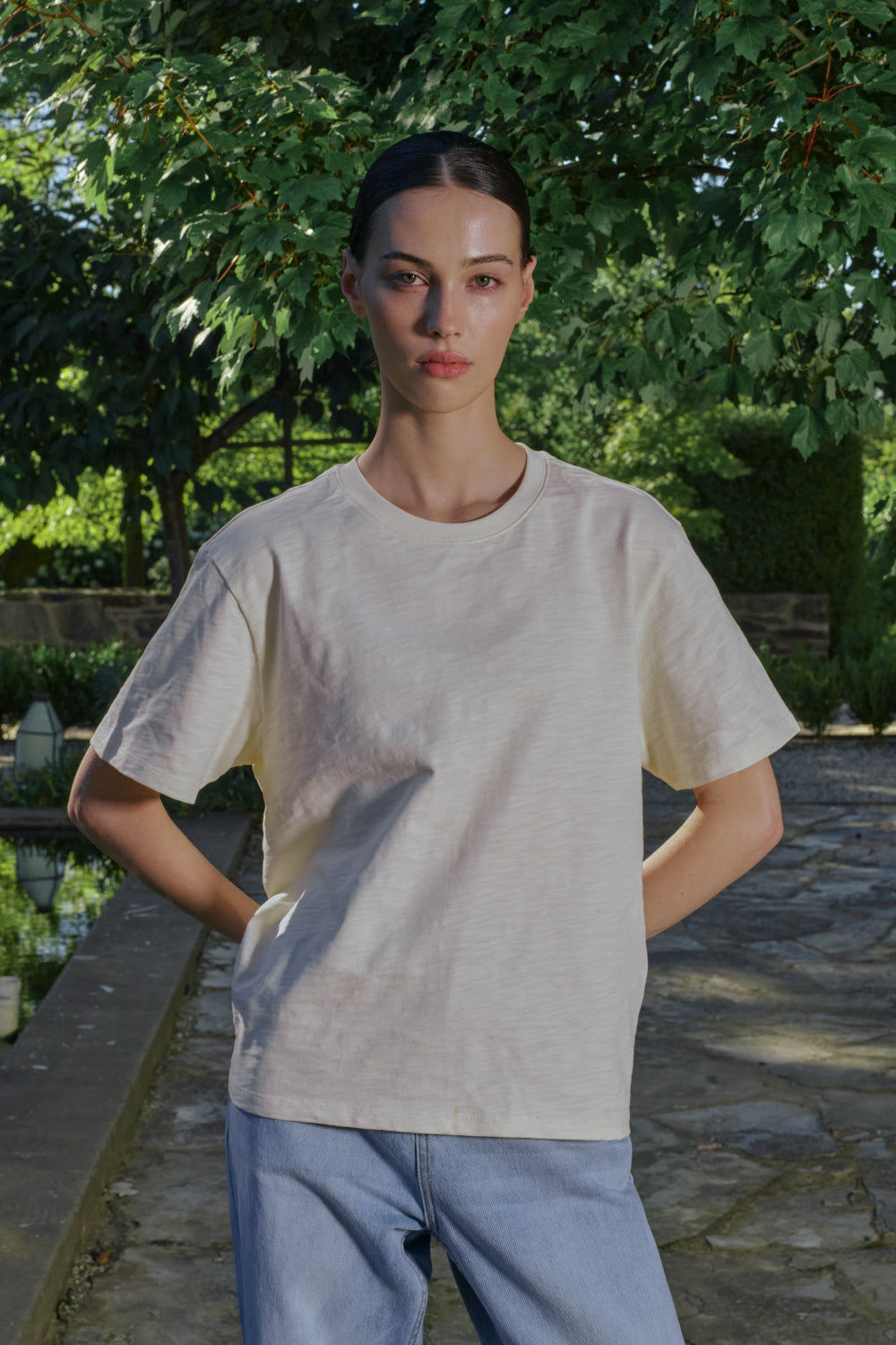 Model wearing a beige t-shirt and light blue pants standing outdoors with greenery in the background