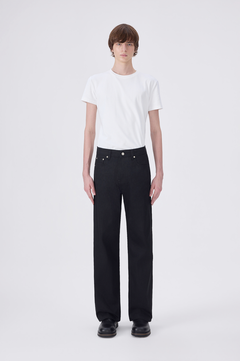 A model stands against a light gray studio background wearing a plain white short-sleeve T-shirt, black straight-leg jeans, and black shoes.