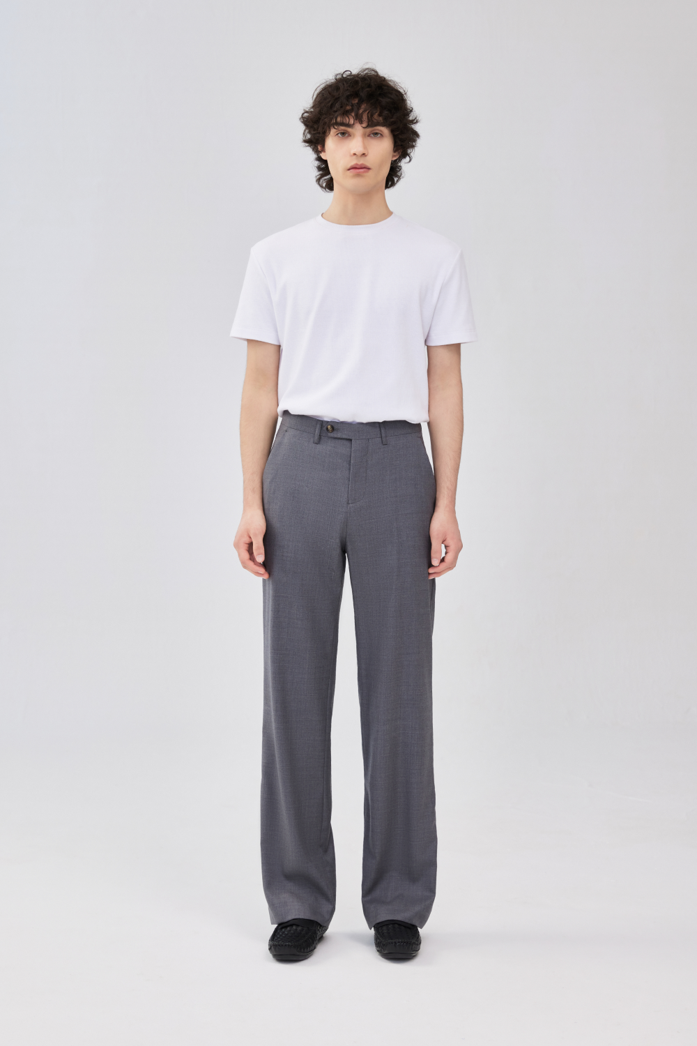 Full-length front view of a model wearing grey wool trousers with a straight leg, front crease, belt loops, side pockets, and button closure, styled with a white T-shirt.