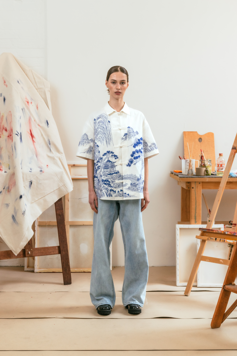 Person wearing a white shirt with blue patterns and light blue jeans in an art studio setting.