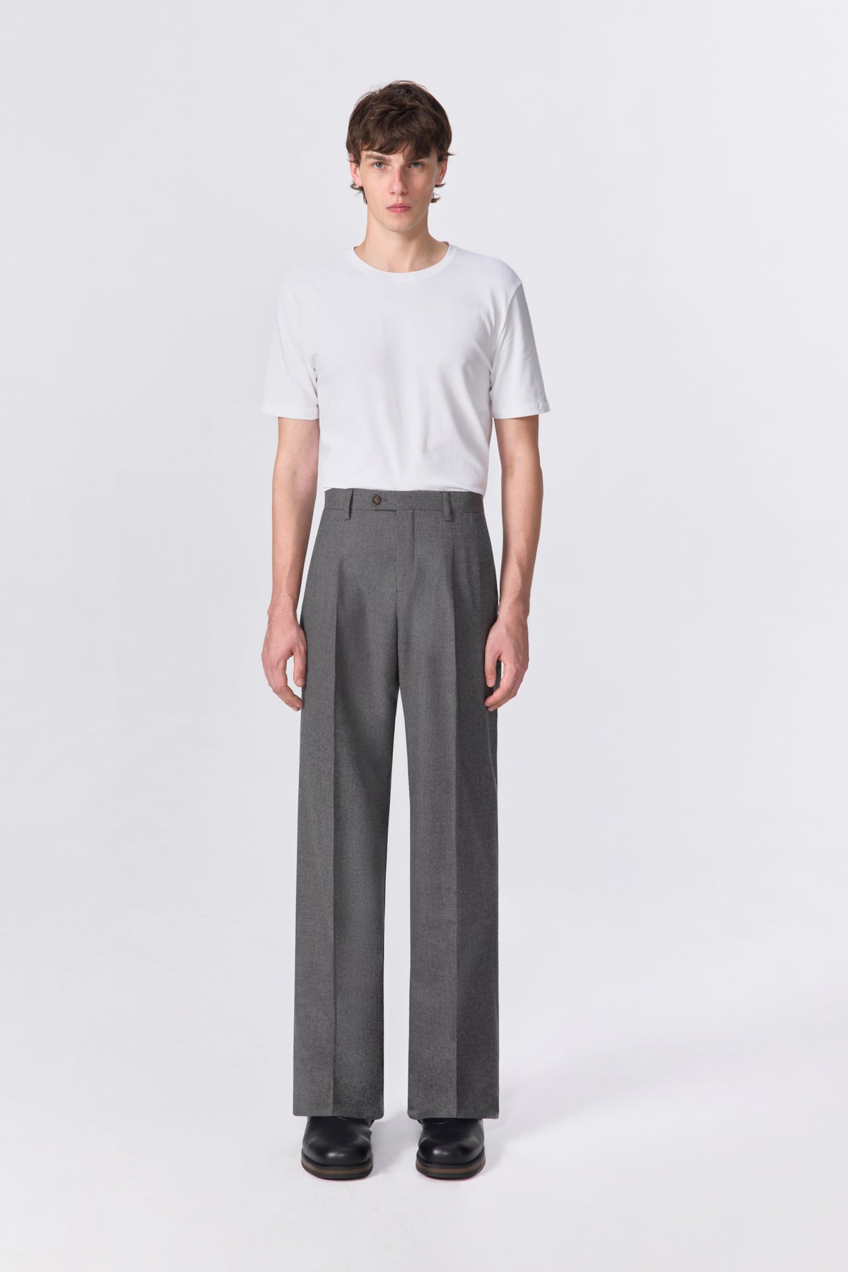 Full-length front view of model wearing grey pleated relaxed straight-leg fit wool trousers.
