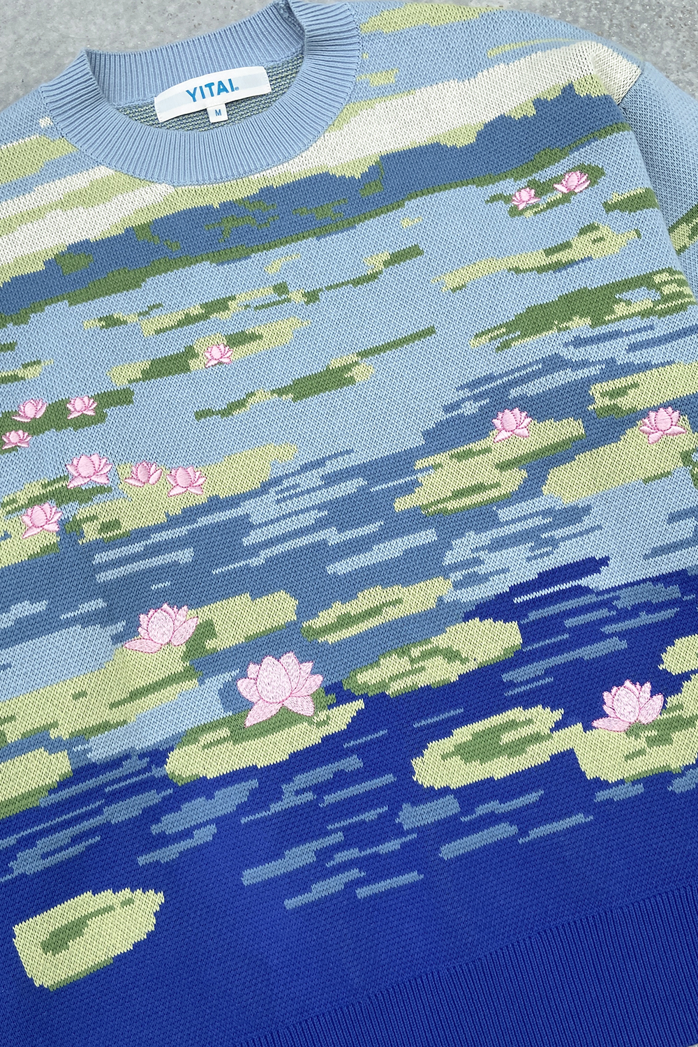 Flat lay of a light blue sweater with a lily pond jacquard knit design, featuring floating lily pads, pink lotus flower embroidery, and deep blue water along the hem.