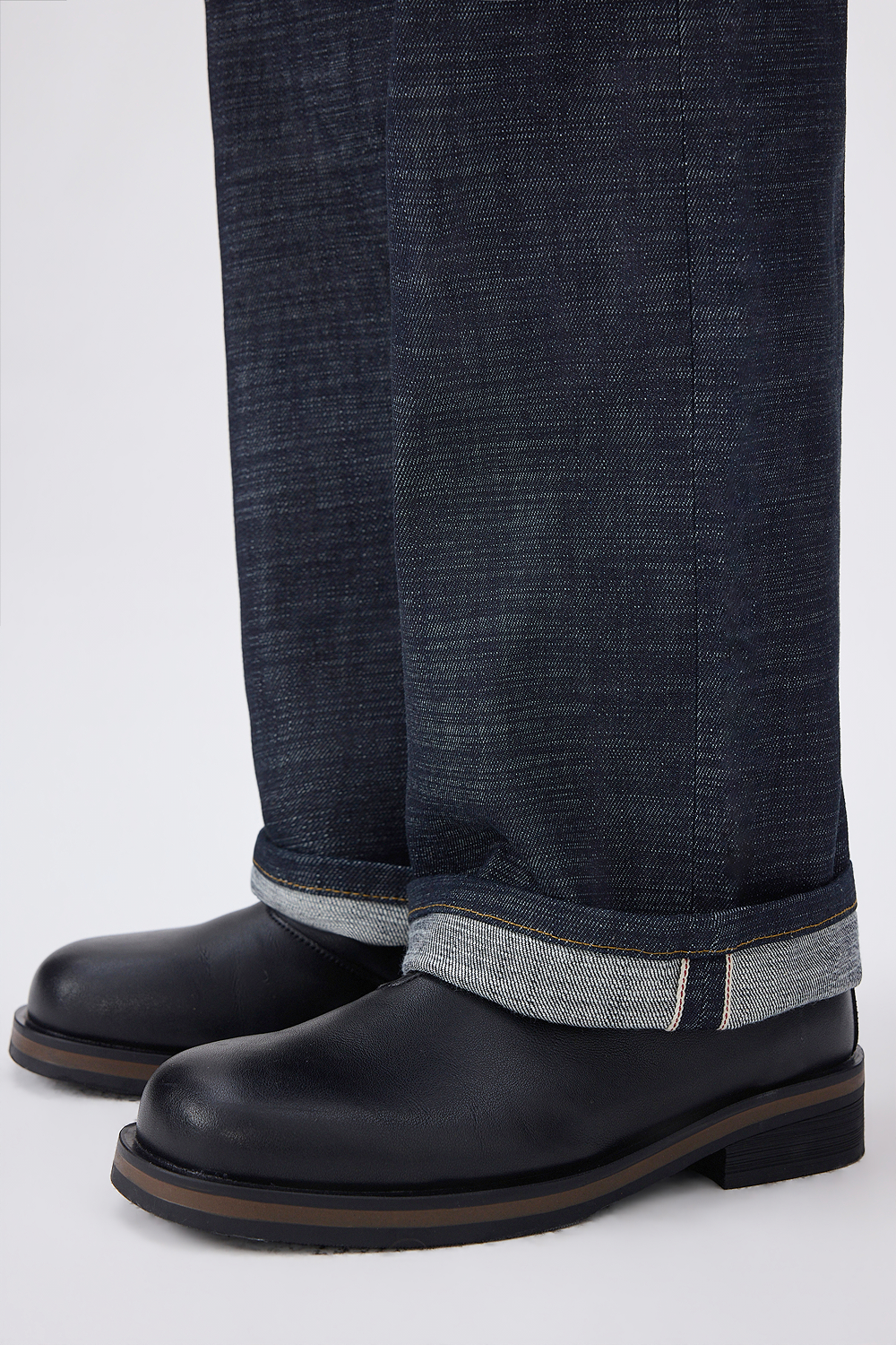 Close-up of cuffed dark indigo selvedge denim jeans showing the selvedge detail and contrast gold stitching, paired with black leather boots.