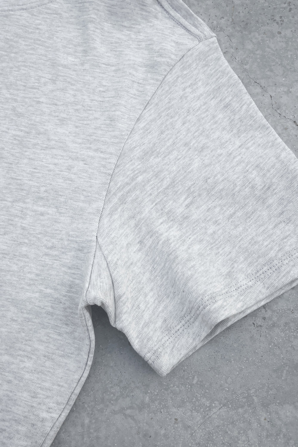 Close-up of the sleeve and side seam of a light heather gray T-shirt laid flat on a gray textured surface, highlighting the soft fabric and stitching details.