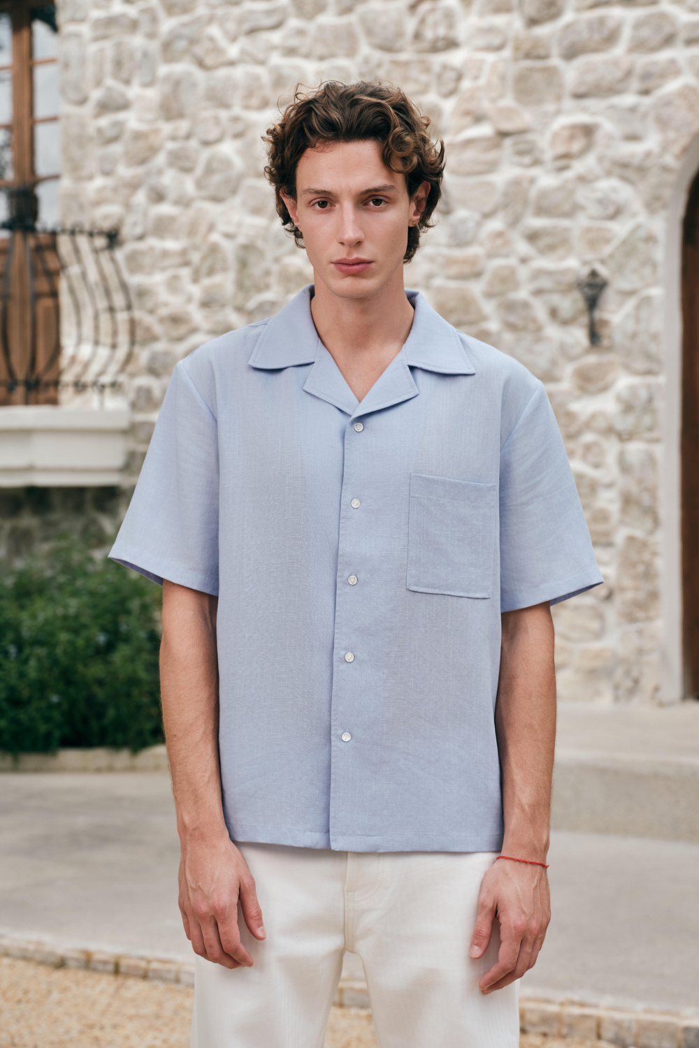 Model wearing a light blue short-sleeve button-front shirt with an open collar and chest patch pocket.