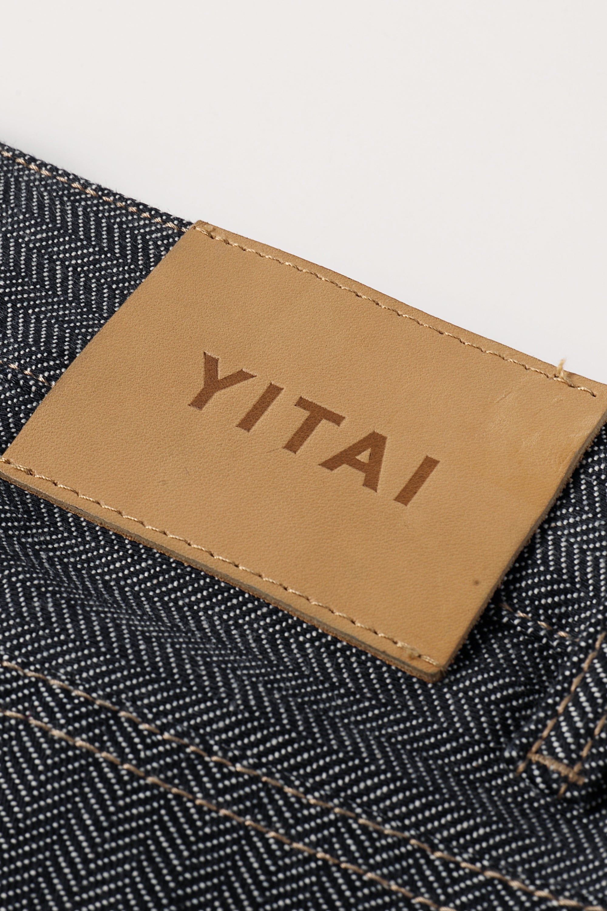 Close-up of a tan leather YITAI brand patch sewn onto a grey herringbone denim fabric.
