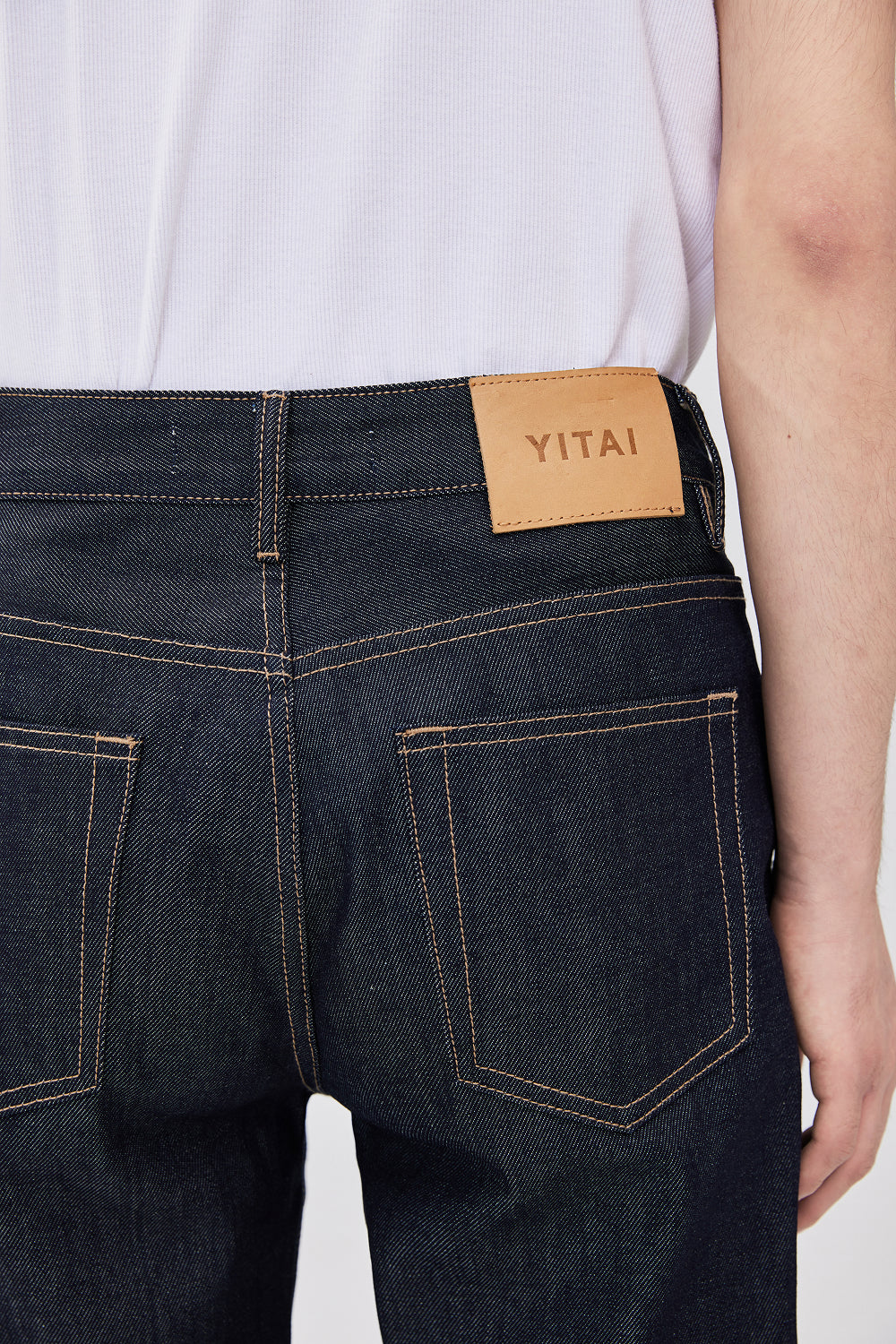 Close-up back view of a model wearing dark indigo denim jeans with contrast stitching and a tan leather YITAI brand patch at the waistband.