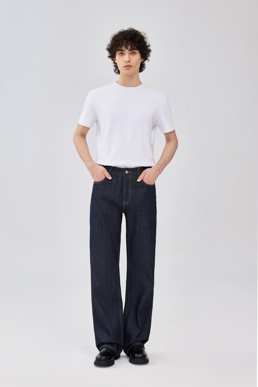 A model wearing indigo blue selvedge denim jeans, a plain white short-sleeve crewneck t-shirt, and black shoes.