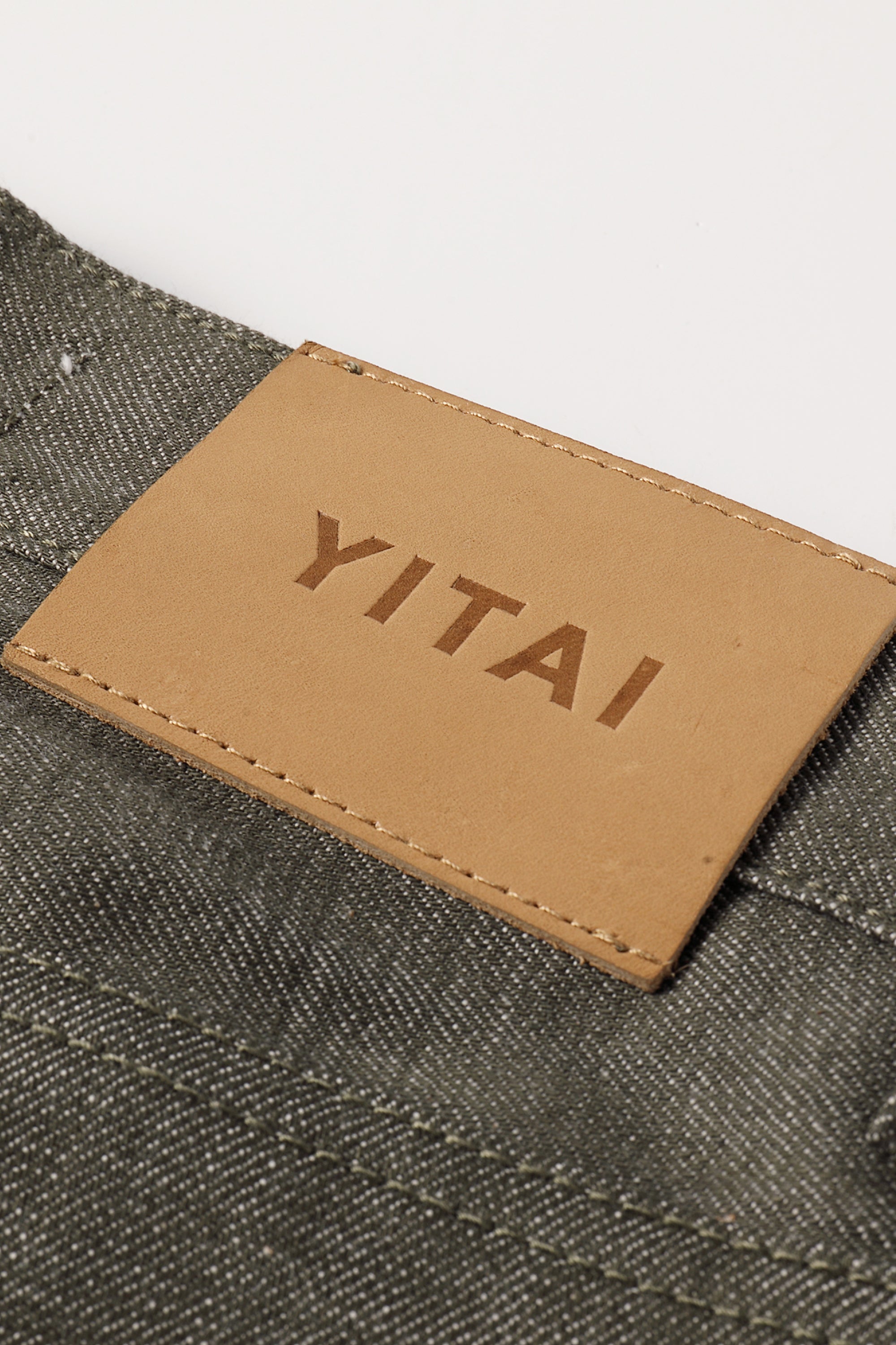 Close-up of a tan leather YITAI brand patch sewn onto a green selvedge denim fabric.