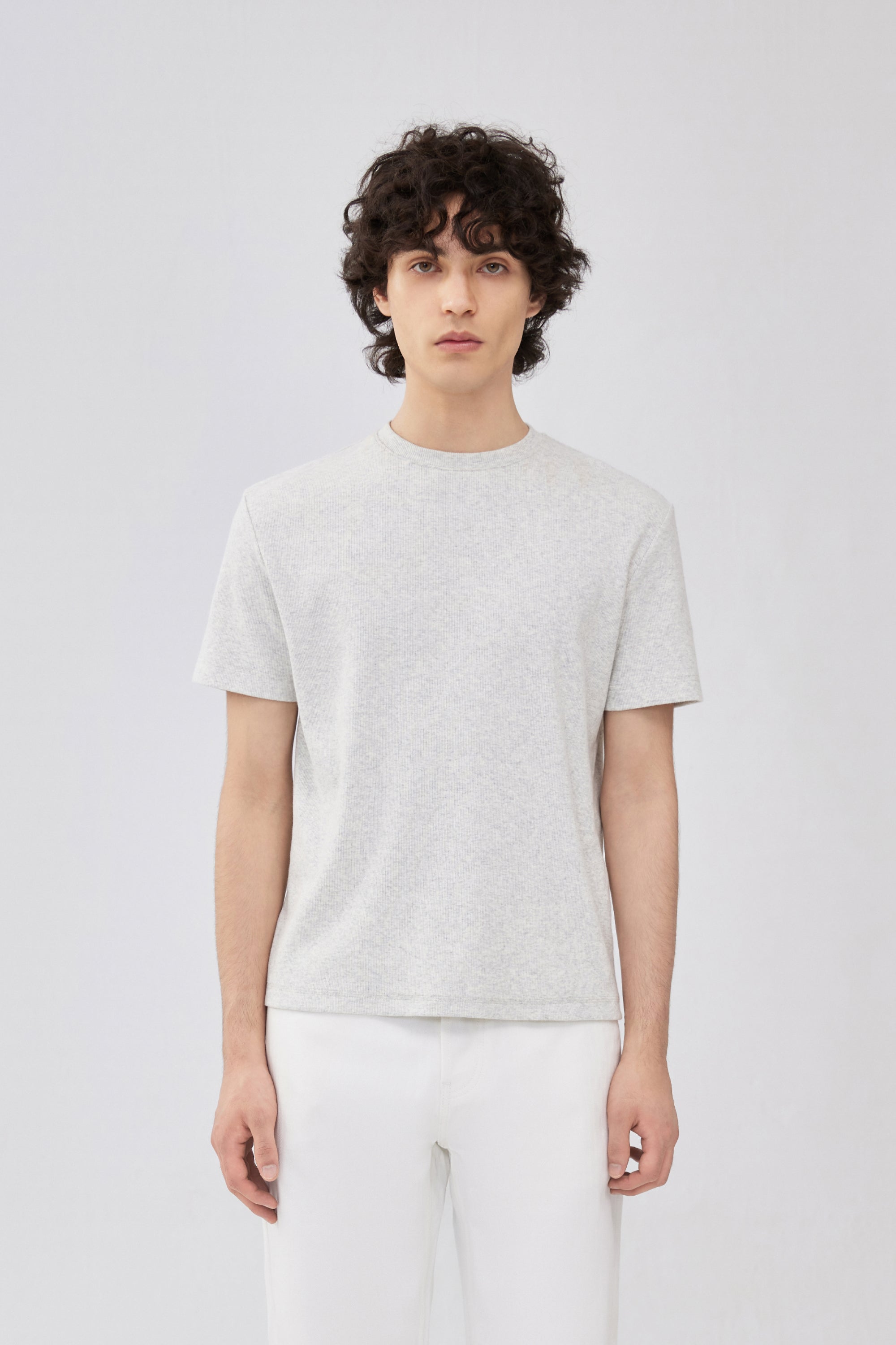 A male model wearing a light grey short-sleeve crewneck t-shirt and white pants, standing against a light gray studio background.