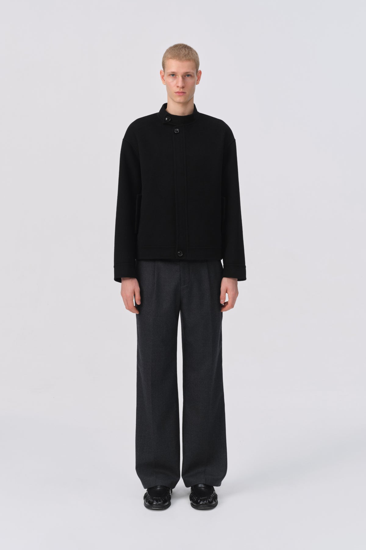 A model stands against a light gray studio background wearing a black cropped button-front jacket with a high collar and dark wide-leg trousers.